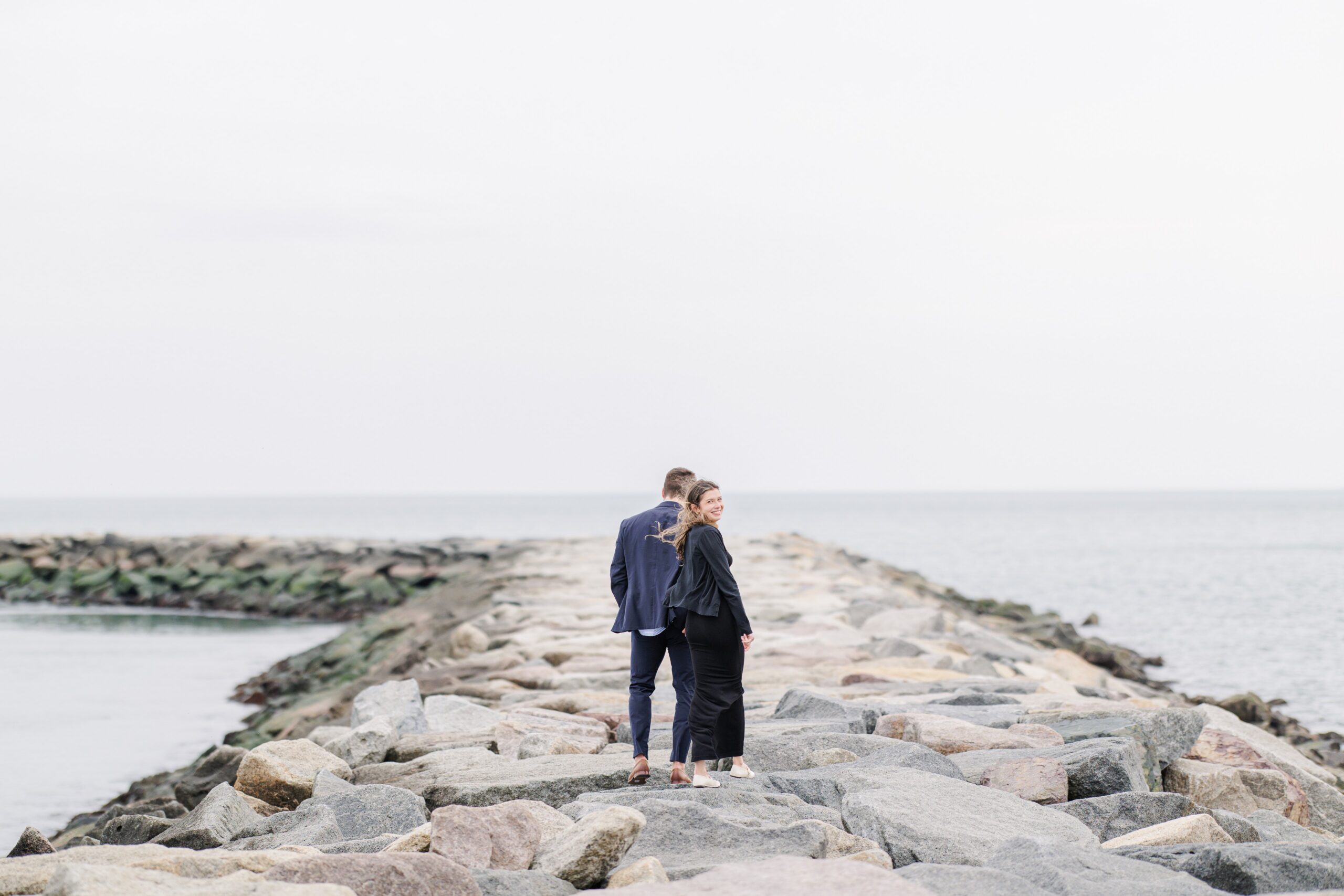 Couple walking away hand in hand on rocky jetty after Scituate Lighthouse proposal in Scituate MA