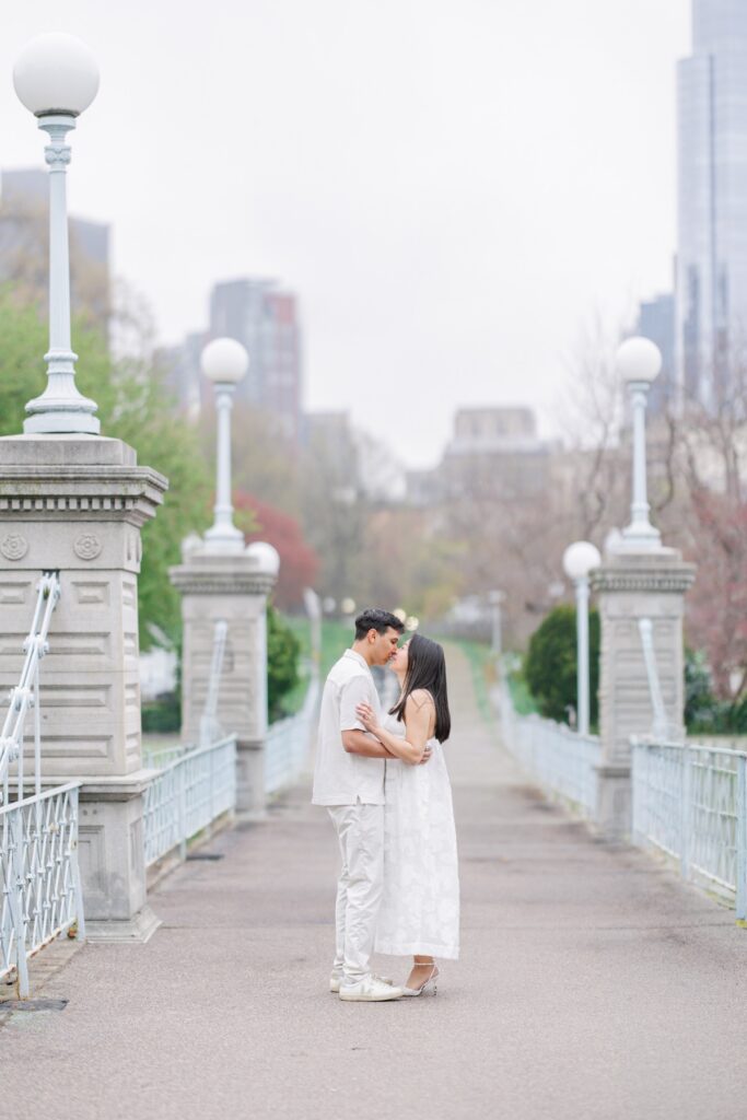     Couple kissing on the bridge in the Boston Public Garden with the Boston skyline behind them during Beacon Hill engagement photos