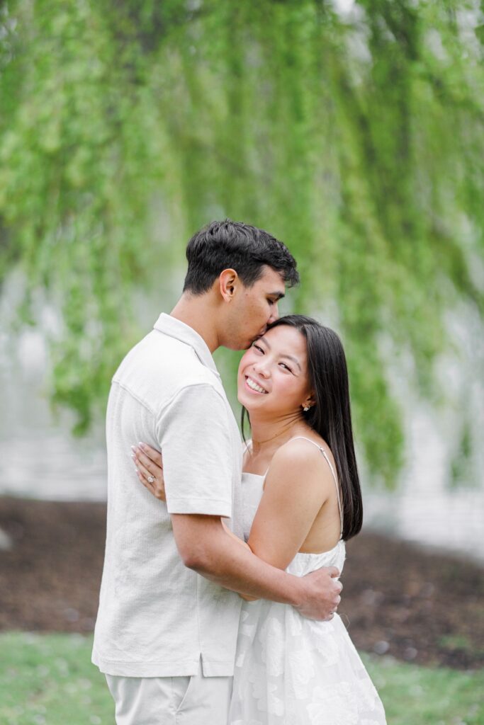     Groom kissing bride’s forehead beneath willow trees in the Boston Public Garden during Beacon Hill engagement photos