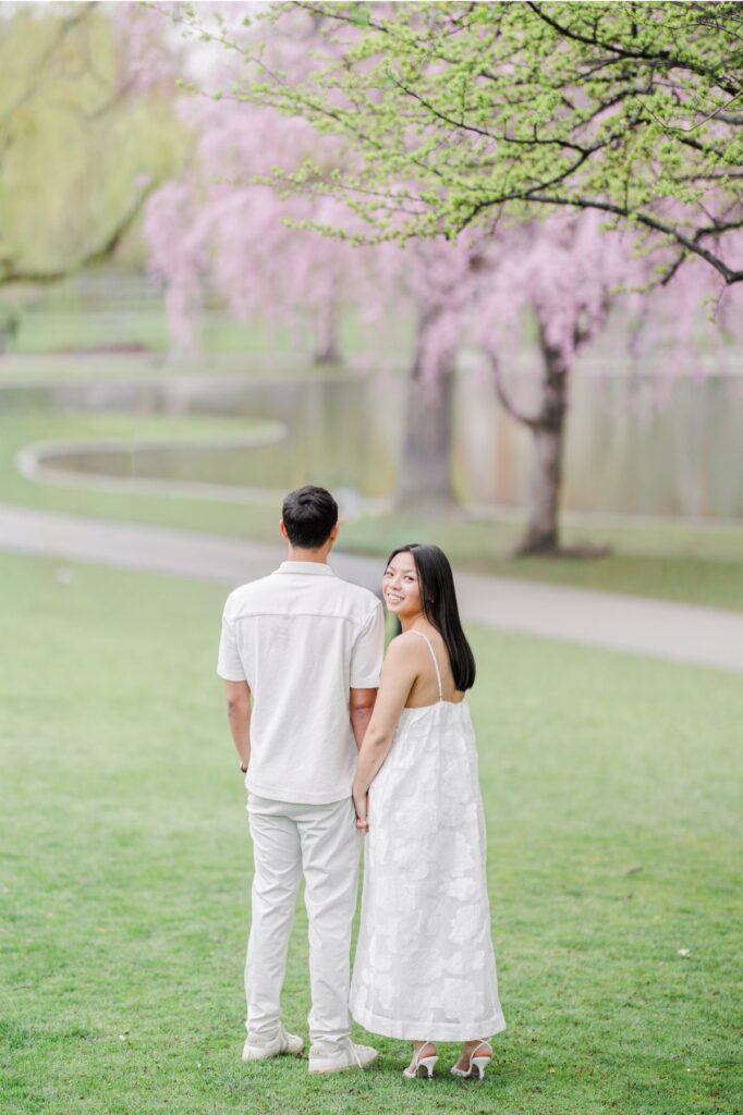     Couple holding hands beneath blooming trees in the Boston Public Garden during a Beacon Hill engagement session