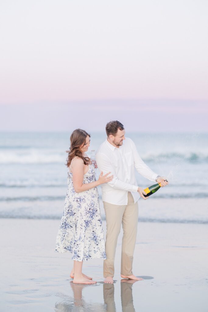     Couple celebrating with champagne during playful New Hampshire engagement photos at sunset in Seabrook, NH.