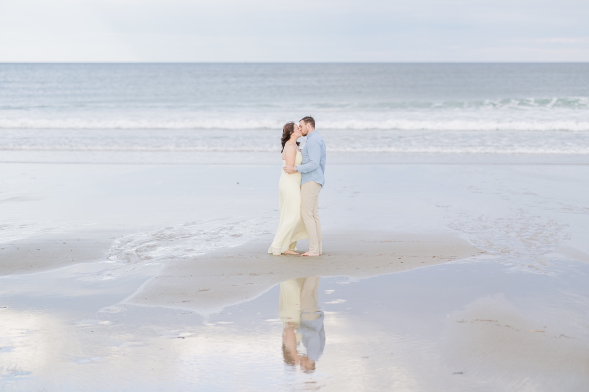Romantic New Hampshire engagement photos of a couple kissing barefoot on the beach with ocean reflections behind them.