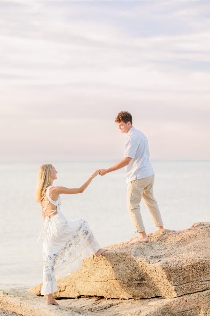 Couple climbing seaside rocks together during a Cape Cod beach engagement session.