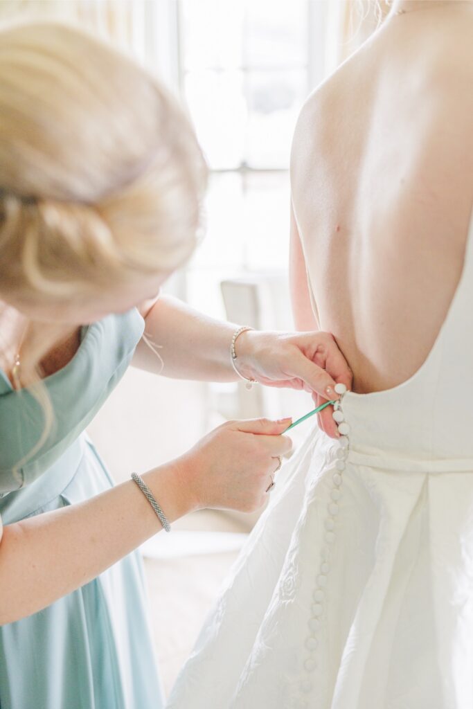 Bridesmaid fastening the buttons on the back of the bride’s wedding gown.