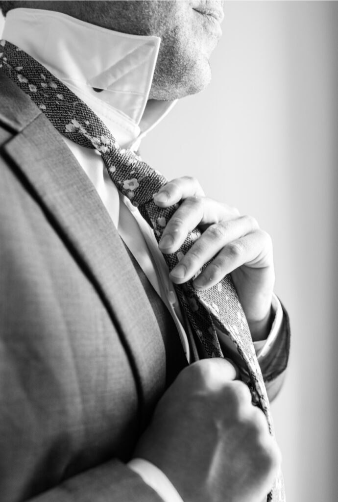 Black-and-white photograph of the groom adjusting his floral necktie.
