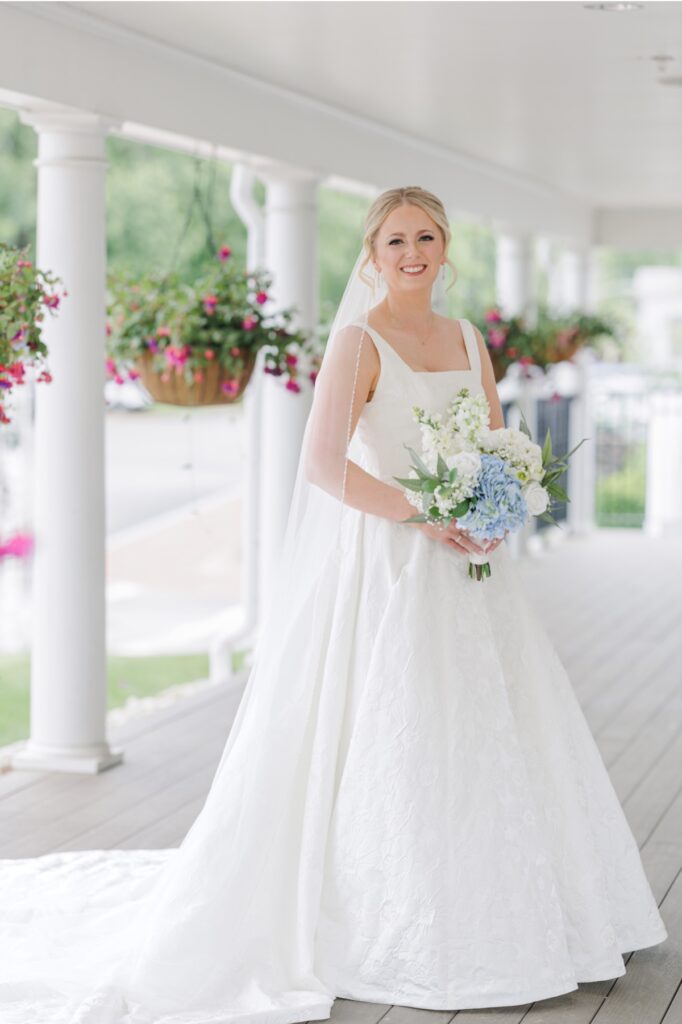 Bride holding a blue hydrangea bouquet on the flower-lined Publick House porch.