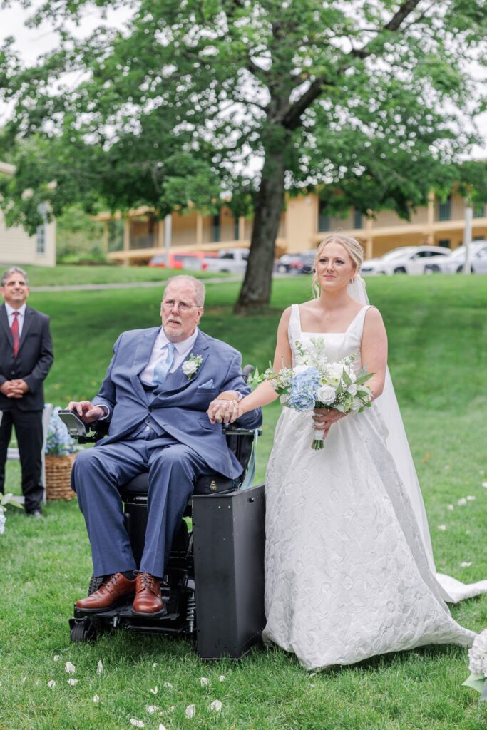 Bride walks beside her father in a power wheelchair during the outdoor wedding ceremony.