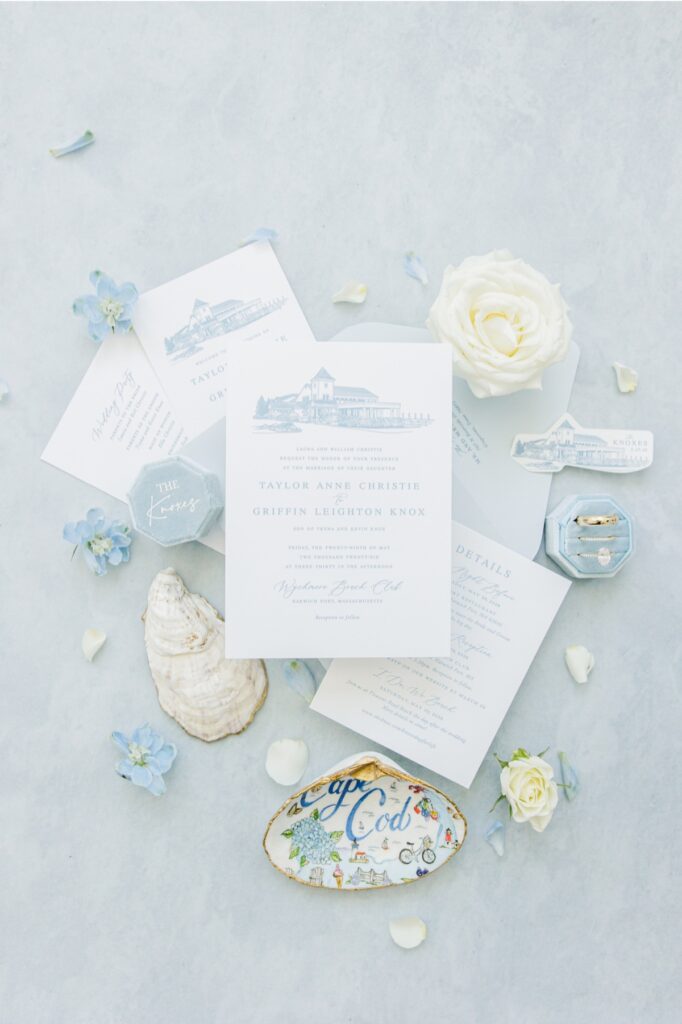 Flat lay of blue and white wedding stationery for a Wychmere Beach Club wedding in Harwich, MA surrounded by white roses, blue flowers, oyster shells, wedding rings, and Cape Cod–inspired details. The invitation features an illustration of Wychmere Beach Club and welcomes guests to the wedding of Taylor Anne Christie and Griffin Leighton Knox.
