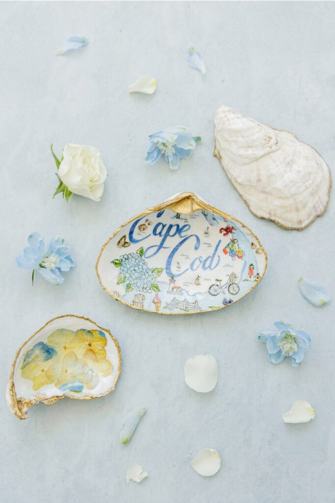 Hand-painted oyster shells with gold edges and Cape Cod illustrations are styled with white roses, blue flowers, and a natural oyster shell on a soft blue backdrop. One shell reads “Cape Cod” and another features pressed hydrangea petals, reflecting coastal wedding details.