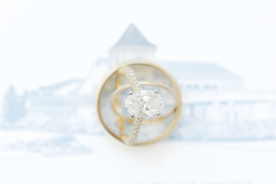 Close-up of gold wedding bands framing an oval diamond engagement ring with the illustrated Wychmere Beach Club invitation softly blurred in the background. The composition highlights the rings against the custom wedding stationery.