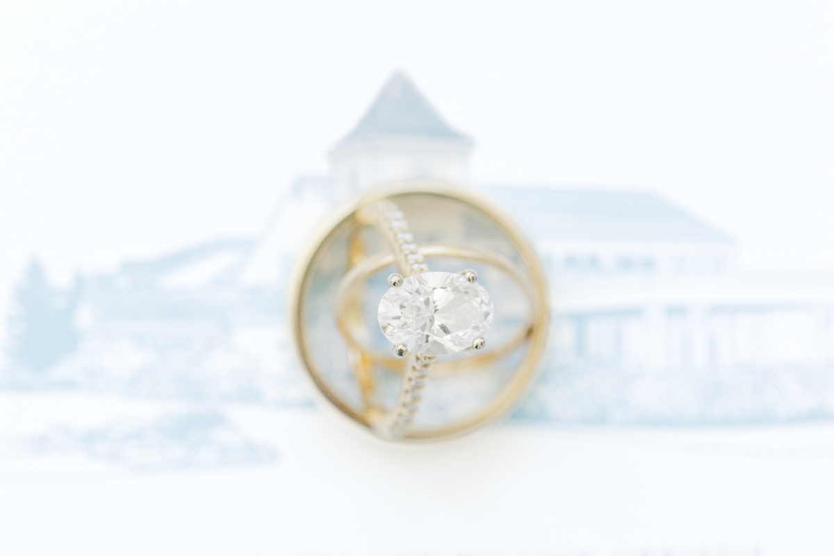Close-up of gold wedding bands framing an oval diamond engagement ring with the illustrated Wychmere Beach Club invitation softly blurred in the background. The composition highlights the rings against the custom wedding stationery.