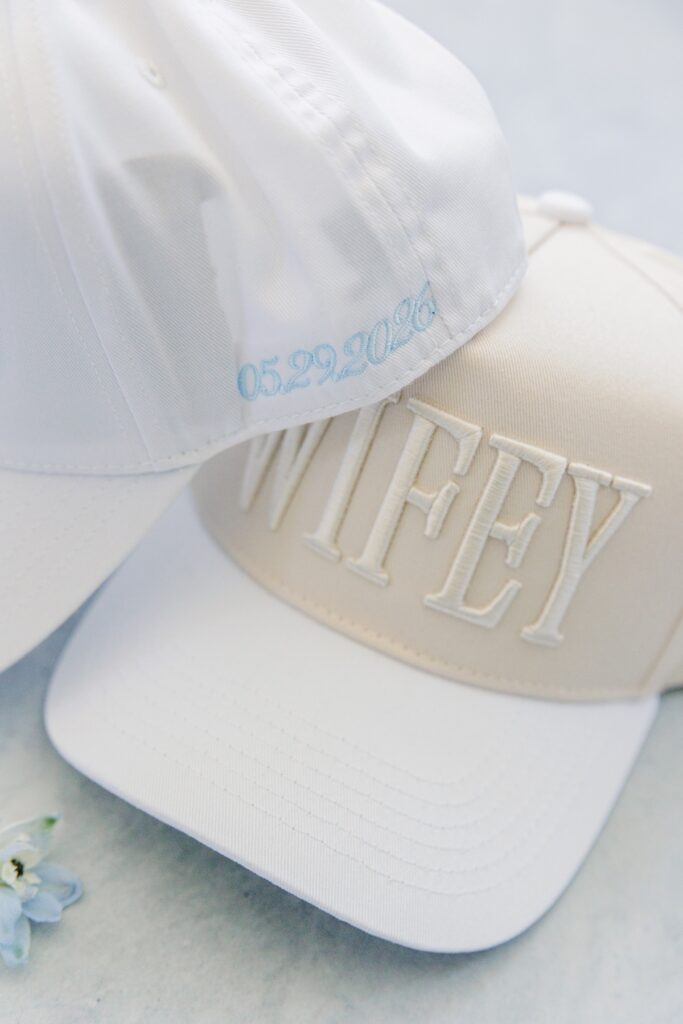 Two white embroidered trucker hats are stacked together with one embroidered “WIFEY” and the other stitched with the wedding date “05.29.2026” in light blue. The personalized accessories are styled as bridal getting-ready details for a Cape Cod wedding.