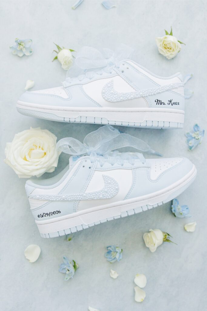 Pair of customized white Nike sneakers with pearl swooshes and sheer ribbon laces displayed among white roses and blue flowers. One shoe is personalized with “Mrs. Knox” and the other with the date “05.29.2026” for a Wychmere Beach Club wedding.