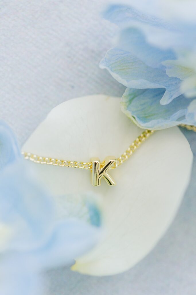 Delicate gold bracelet with the initial “K” rests on a white rose petal surrounded by soft blue flowers. The personalized jewelry complements the bride’s wedding day accessories.