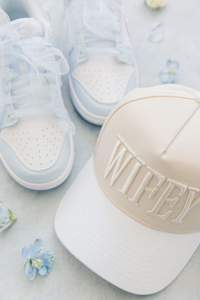 White embroidered “WIFEY” trucker hat is styled beside customized white bridal sneakers with sheer ribbon laces and pearl details. The personalized accessories are photographed as modern bridal details for a Harwich MA wedding.