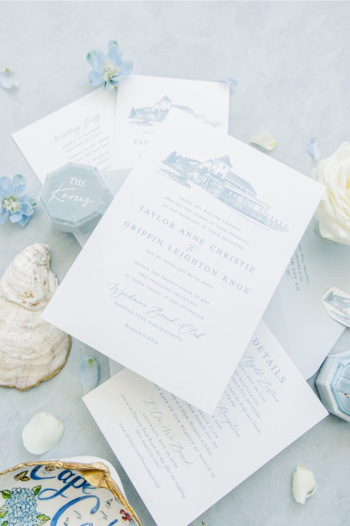 Blue and white wedding invitation suite displayed with oyster shells, blue flowers, white roses, and a blue velvet ring box for a Wychmere Beach Club wedding. The invitation announces the marriage of Taylor Anne Christie and Griffin Leighton Knox at Wychmere Beach Club in Harwich Port, Massachusetts.