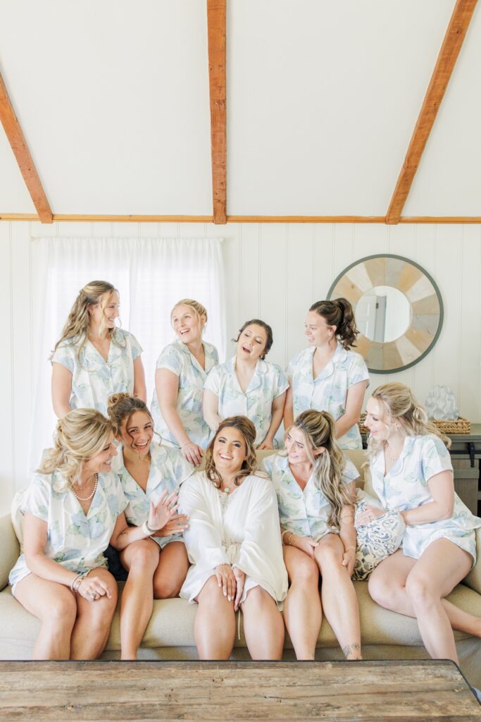 Bride sits surrounded by her bridesmaids in matching blue and white floral pajamas as they laugh together before a Wychmere Beach Club wedding in Harwich, MA. The bright room and coordinated attire capture the excitement of the morning preparations.
