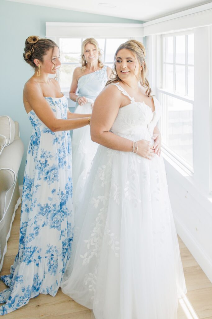 The bride smiles as her sister fastens the back of her wedding dress while her mother looks on beside a sunlit window. The quiet getting-ready moment took place before a Cape Cod wedding celebration.