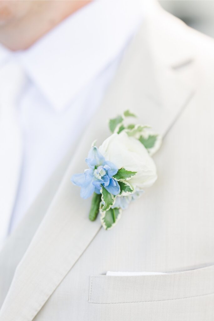 Close-up of the groom’s white rose and blue delphinium boutonniere pinned to the lapel of a light tan suit jacket. The floral design complements the blue and white color palette of the Cape Cod wedding.
