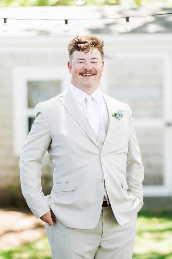 Groom smiles outdoors wearing a light tan suit with a white tie and blue and white boutonniere before the ceremony. The portrait captures him waiting for the start of his Wychmere Beach Club wedding.