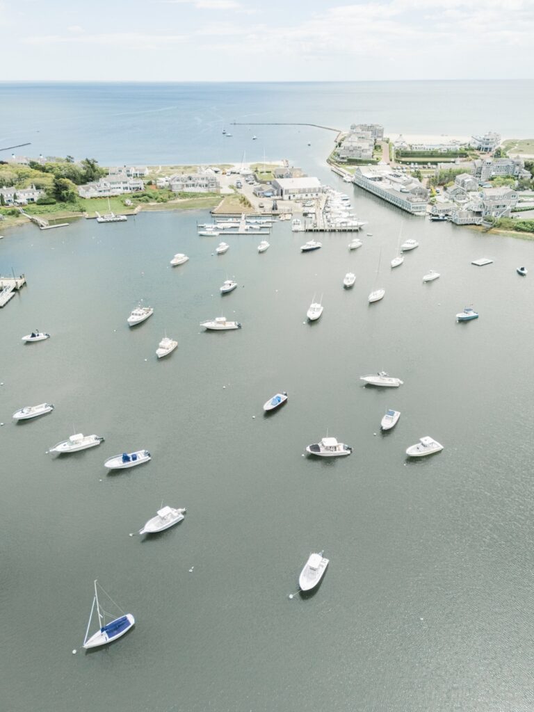 Aerial view of Wychmere Harbor in Harwich Port with sailboats and motorboats anchored across the water on a sunny day. The coastal landscape showcases the waterfront setting of a Wychmere Beach Club wedding on Cape Cod.