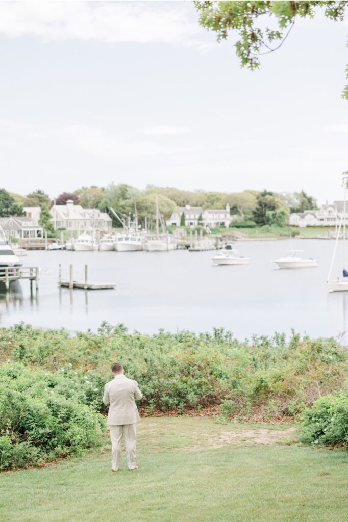 The groom stands with his back turned overlooking Wychmere Harbor before the couple’s first look. Boats and waterfront homes create a classic Cape Cod backdrop for the Wychmere Beach Club wedding.