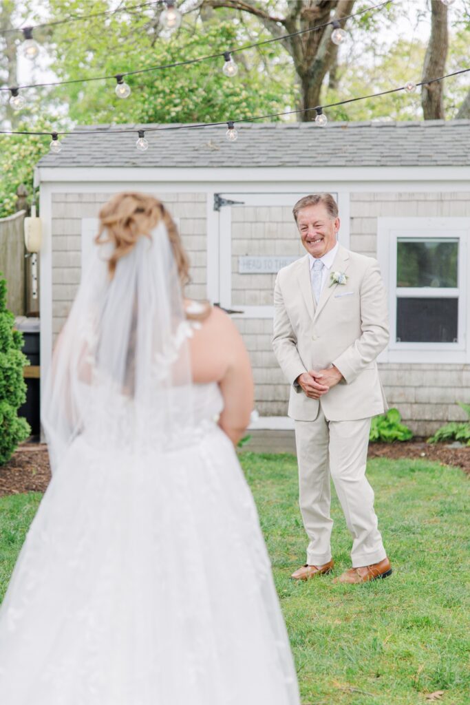 Father of the bride smiles as he turns to see his daughter in her wedding dress for the first time. The emotional first look takes place beneath café lights in a garden before the ceremony.