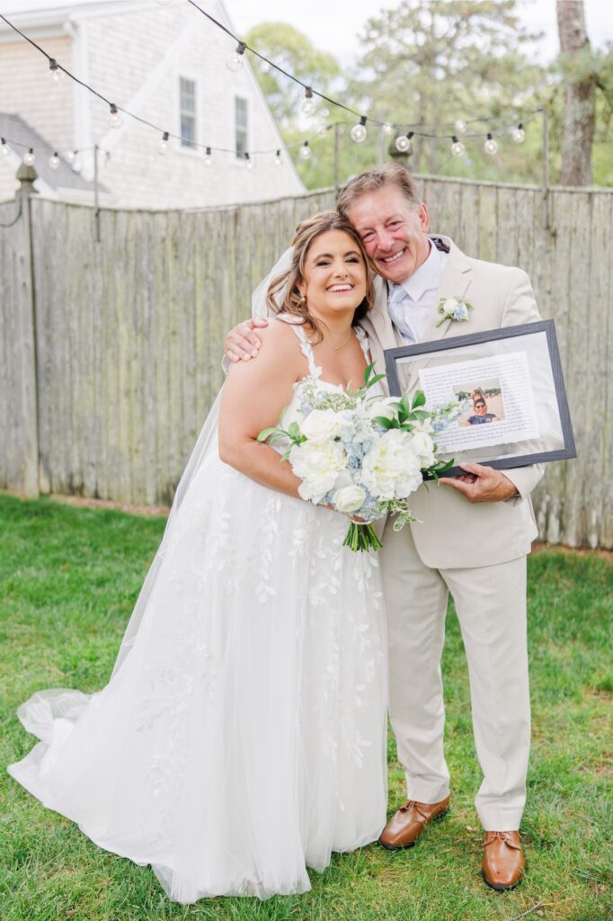 The bride and her father smile together while holding a framed photo gift and a bouquet of white roses, hydrangeas, and blue delphinium. The portrait captures a special moment before their Wychmere Beach Club wedding.