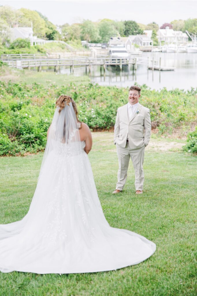 The groom smiles as he turns to see the bride for the first time during their first look at Wychmere Beach Club. The harbor and boats create a scenic waterfront backdrop behind the couple.