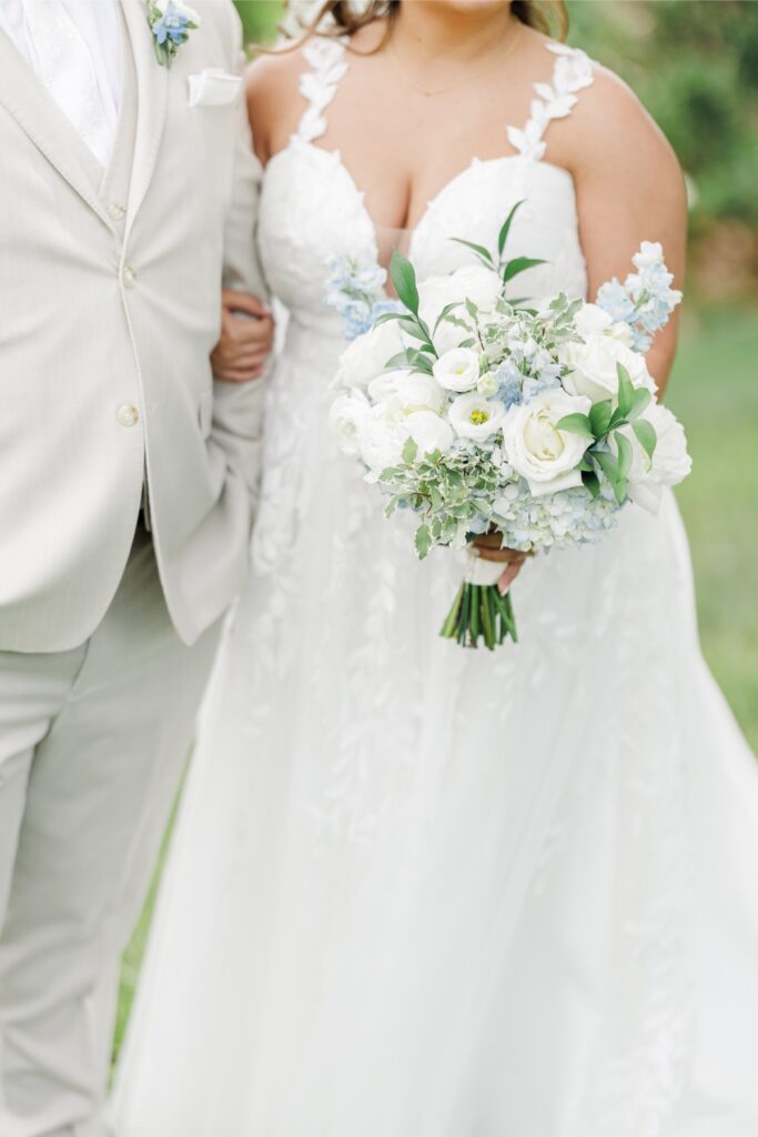The bride holds a bouquet of white roses, hydrangeas, ranunculus, pale blue delphinium, and fresh greenery while walking arm in arm with the groom. The floral arrangement complements the elegant style of their Harwich MA wedding.