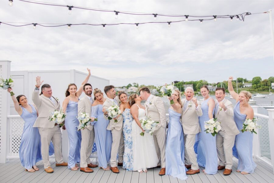 The wedding party celebrates on the waterfront deck at Wychmere Beach Club with the bride and groom sharing a kiss in the center. Light blue bridesmaid dresses and neutral suits reflect the coastal style of the Cape Cod wedding.