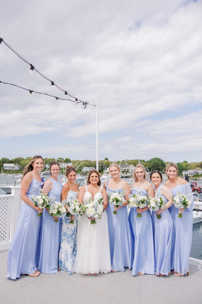 The bride poses with her bridesmaids on the waterfront deck at Wychmere Beach Club. Soft blue dresses and white floral bouquets complement the harbor views behind the group.