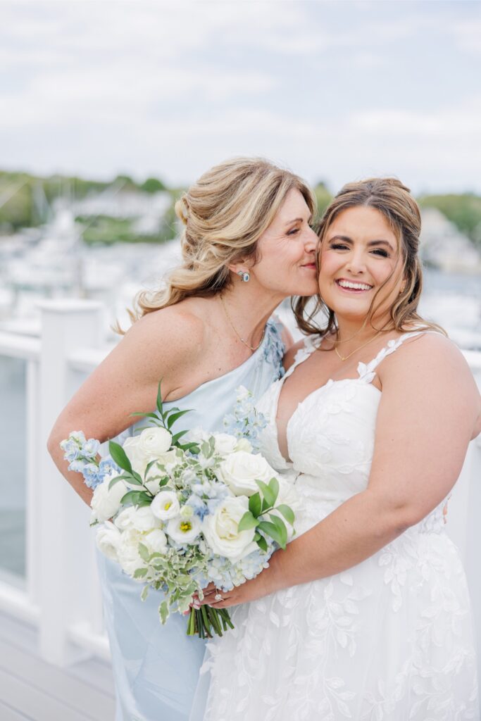 The mother of the bride kisses her daughter’s cheek as they smile together on the waterfront deck at Wychmere Beach Club. The portrait captures a joyful family moment before the ceremony with harbor views in the background.