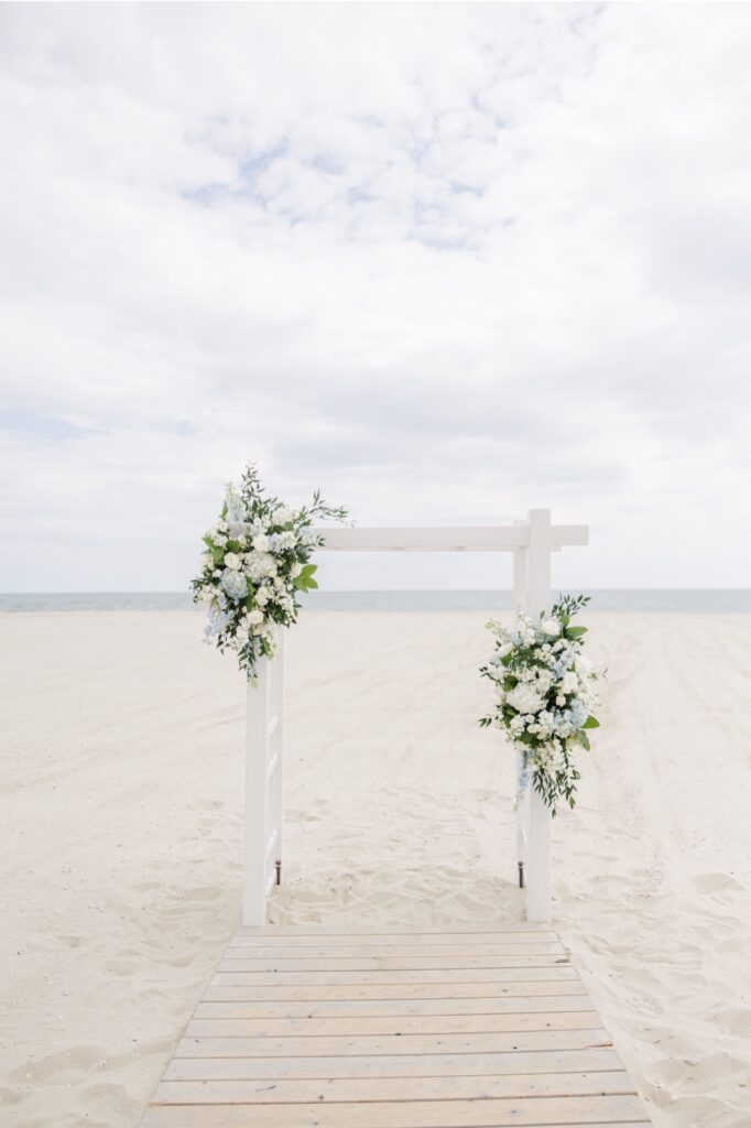 A white wooden wedding arbor decorated with white and pale blue floral arrangements stands on the beach overlooking the ocean at Wychmere Beach Club. The simple ceremony backdrop highlights the coastal setting of the Cape Cod wedding.