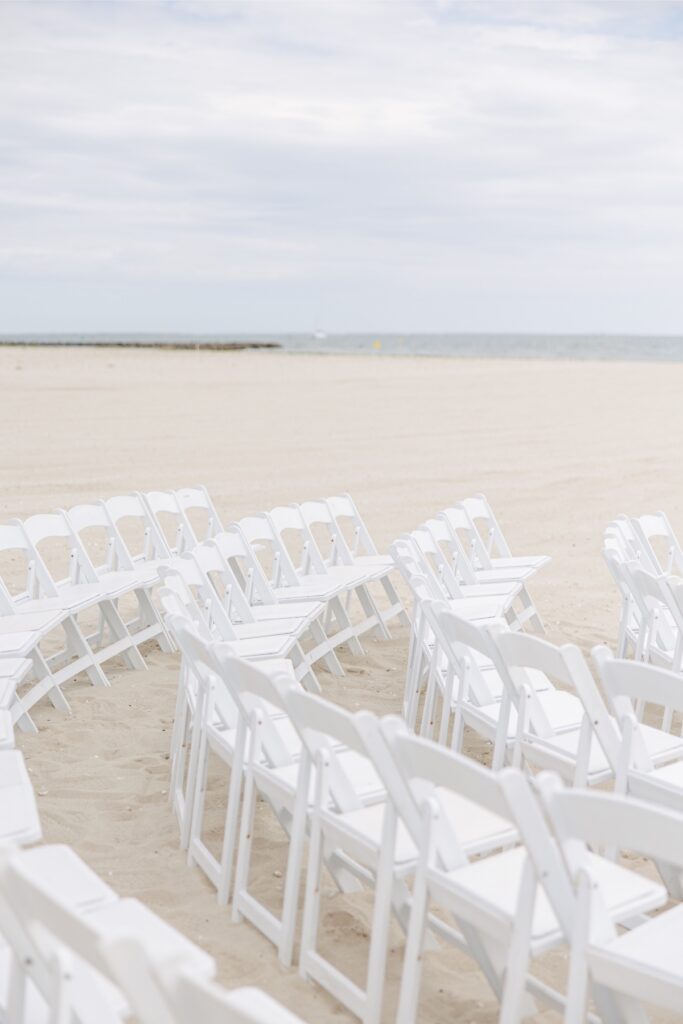 Rows of white folding chairs are arranged on the sand facing the ocean for a beach wedding ceremony at Wychmere Beach Club. The intimate ceremony setup overlooks the Cape Cod shoreline beneath a cloudy sky.