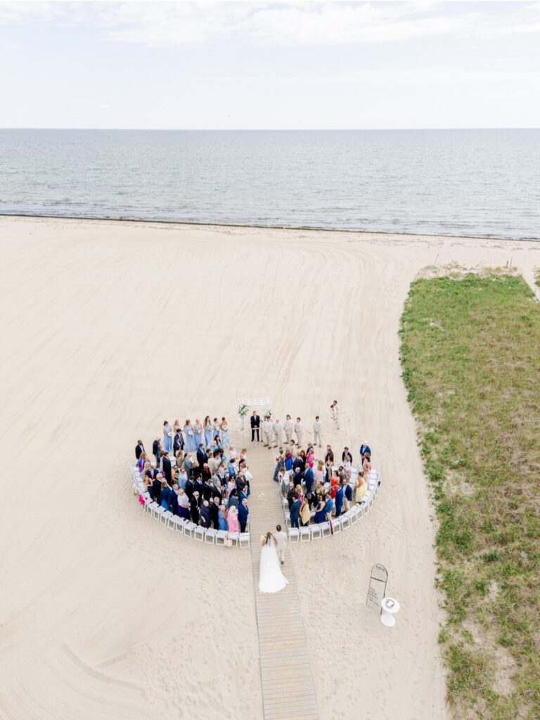 An aerial view captures guests gathered for a beach wedding ceremony at Wychmere Beach Club with the ocean stretching behind them. A wooden walkway leads to the floral ceremony arbor surrounded by family and friends on the sand.