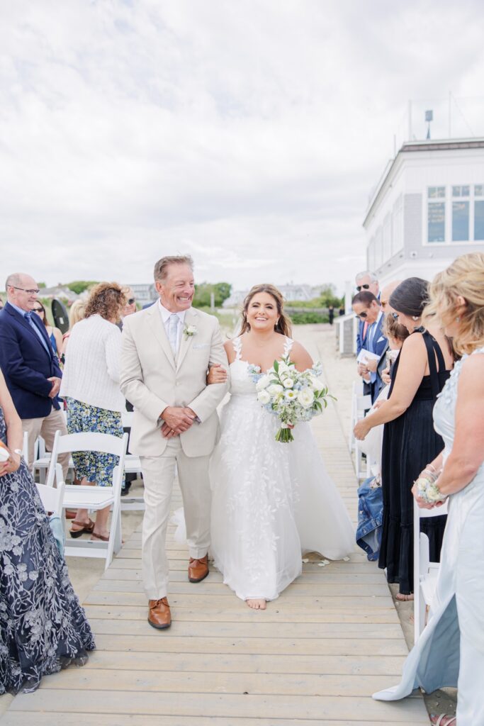 The bride walks down the wooden aisle with her father toward the beach ceremony at Wychmere Beach Club. Guests stand on either side as she carries a bouquet of white and pale blue flowers.
