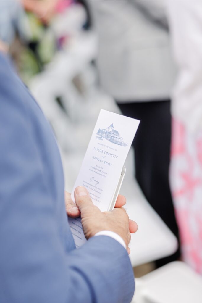A wedding guest holds the ceremony program while seated before the beach ceremony at Wychmere Beach Club. The printed program features the couple’s names and ceremony details in a classic blue design.
