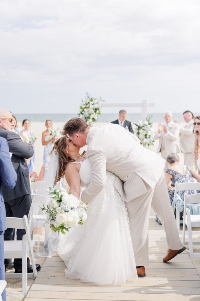 The bride and groom share their first kiss as newlyweds during their beach ceremony at Wychmere Beach Club. Family and friends applaud as the couple celebrates against the Cape Cod ocean backdrop.