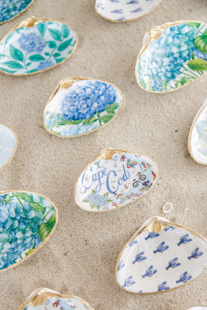 Hand painted oyster shells with blue hydrangea patterns, Cape Cod illustrations, and gold edges are displayed on the sand as coastal wedding details. The personalized shells reflect the seaside style of the Wychmere Beach Club wedding.