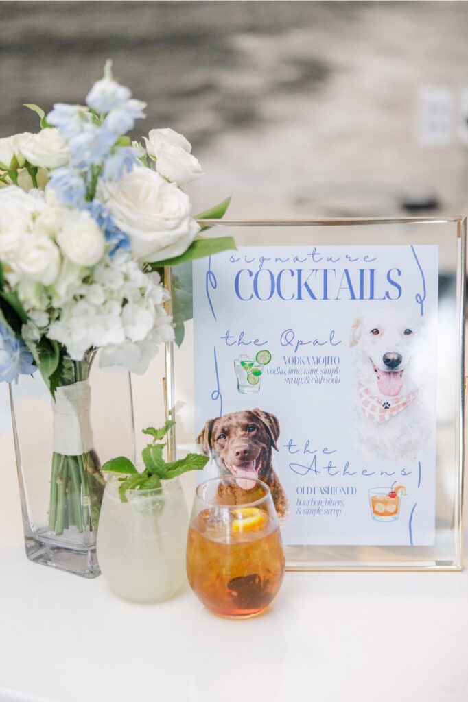 A wedding signature cocktail display features a bouquet of white and pale blue flowers alongside two drinks and a custom sign illustrated with the couple’s dogs. The sign reads “signature COCKTAILS.” “the Opal. VODKA MOJITO. vodka, mint, simple syrup, & club soda.” “the Athens. OLD FASHIONED. bourbon, bitters, & simple syrup.”