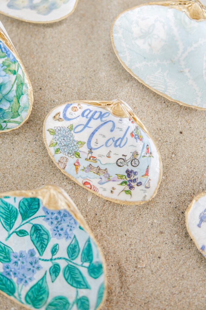 A hand painted oyster shell with gold edging rests on the sand surrounded by other decorative shells. The shell features “Cape Cod” with watercolor illustrations of hydrangeas, lighthouses, sailboats, bicycles, and other coastal landmarks.