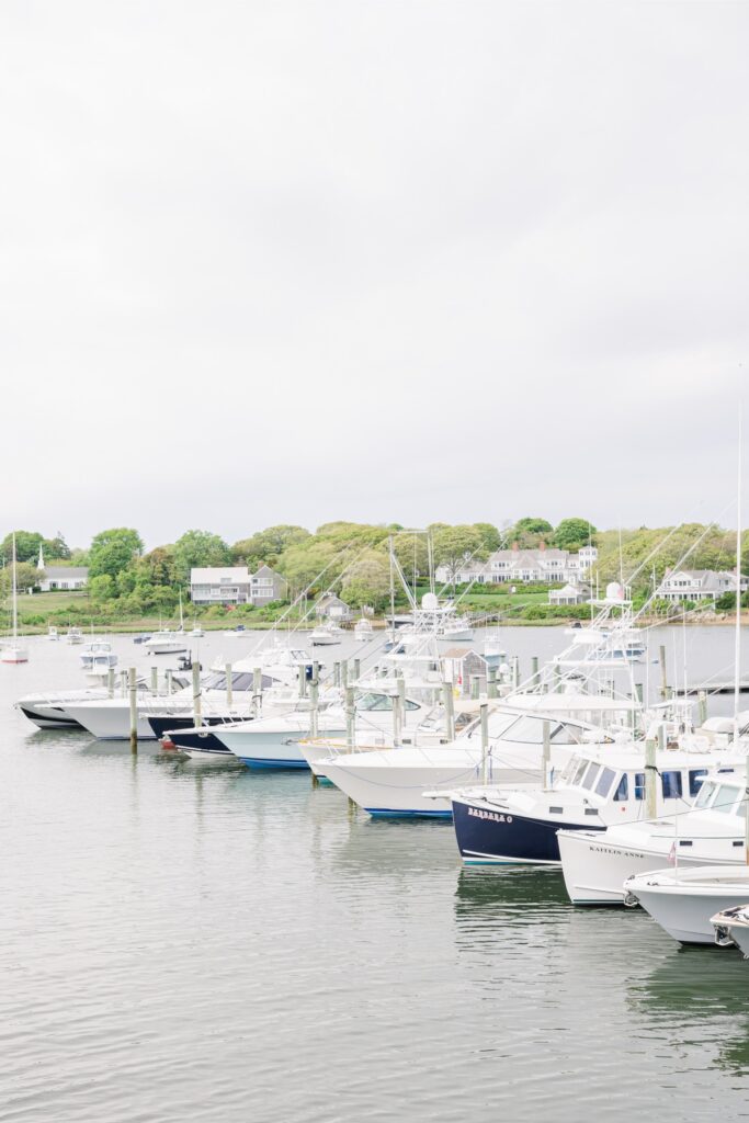 Boats are docked in a Cape Cod marina with waterfront homes and green shoreline in the background. The calm harbor provides a classic coastal view near the wedding venue.