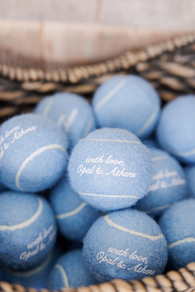 A basket filled with pale blue tennis balls serves as a wedding favor inspired by the couple’s dogs. The tennis balls are printed with “with love, Opal & Athens.”