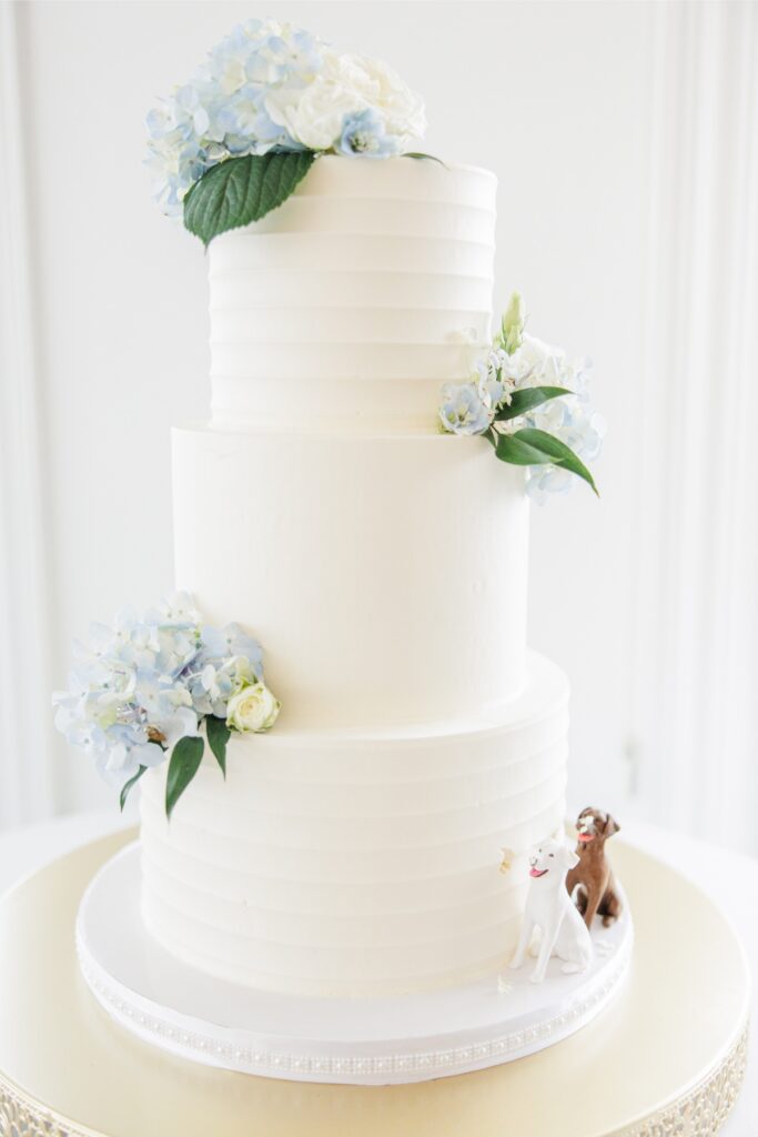 A three tier white wedding cake is decorated with white roses, pale blue hydrangeas, and greenery cascading between the tiers. Small dog figurines at the base add a personalized tribute to the couple’s pets.