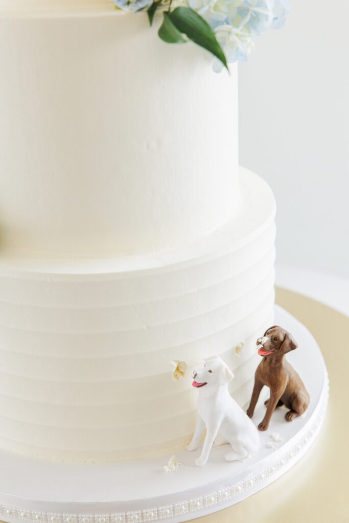 Close up of the base of a white wedding cake featuring two dog figurines beside small bite marks in the frosting. The playful cake detail celebrates the couple’s dogs as part of the wedding design.