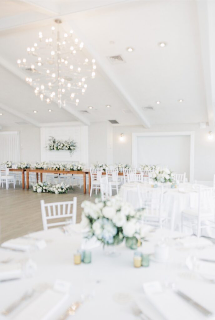 A bright reception room is set with round guest tables, long head tables, and white floral arrangements beneath modern chandeliers. The airy space is ready for guests to enjoy the wedding reception.
