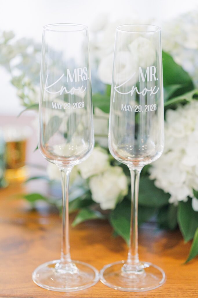 Two champagne flutes are engraved with “MRS. Knox. MAY 29, 2026.” and “MR. Knox. MAY 29, 2026.” The personalized glasses are displayed in front of white and pale blue wedding flowers.