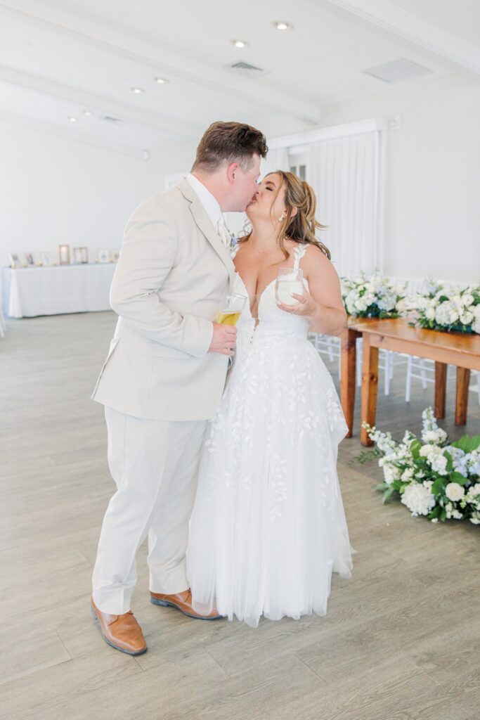 The bride and groom share a kiss while holding drinks inside their wedding reception space. White floral arrangements and the head table create an elegant backdrop for the candid celebration.