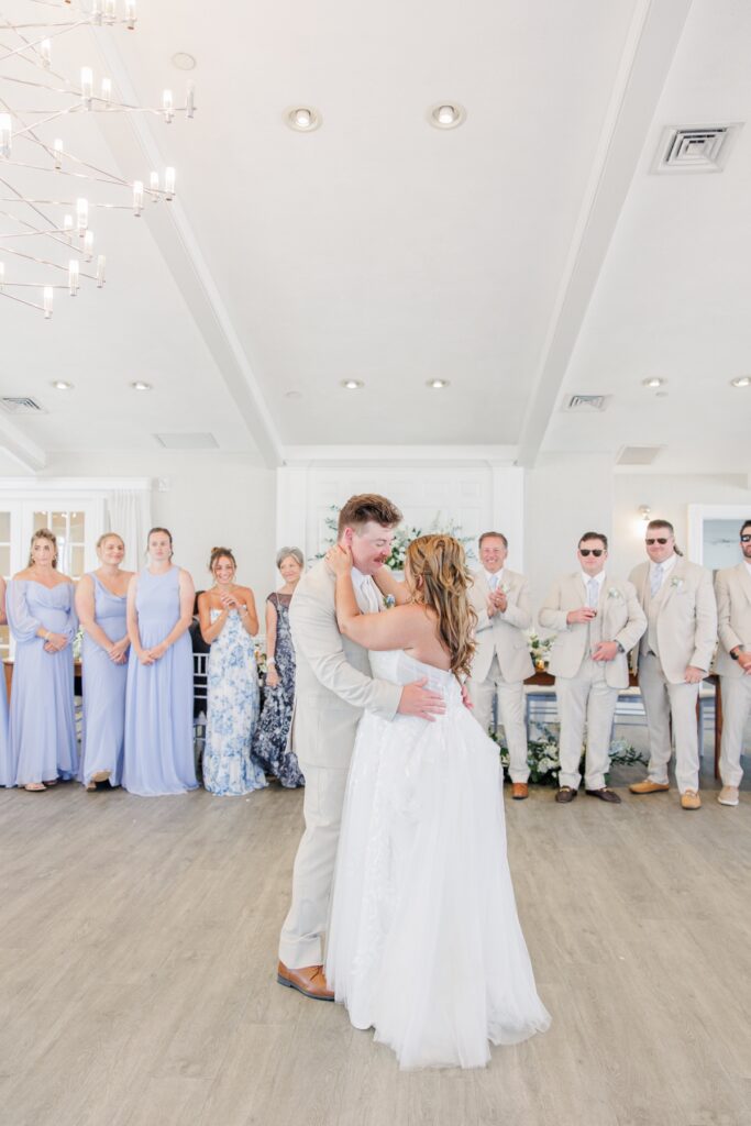 The bride and groom share their first dance while family and wedding party members watch with smiles around the dance floor. The joyful moment captures the couple surrounded by their closest loved ones.